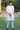 A man wearing a light-colored traditional kurta and trousers standing outdoors on a grassy lawn with trees and a stone structure in the background.