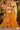 A woman models a bright yellow and orange printed lehenga choli with a matching dupatta in a decorative indoor set. An inset shows a closer view of the choli.