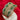 A wide, ornate gold-toned bangle bracelet with green and silver pavé stone detailing, displayed on a white round stand over a red velvet background. A small white tag hangs from the bracelet.