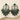 A pair of ornate silver-toned statement earrings with deep green gemstone accents and dangling teardrop charms, displayed on a beige wavy surface with dried floral props. A small label reading No-50 appears in the top right corner.