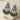 A pair of ornate green and silver dangle earrings displayed on a cream-colored star-shaped stand, with dried decorative grass in the lower left corner.